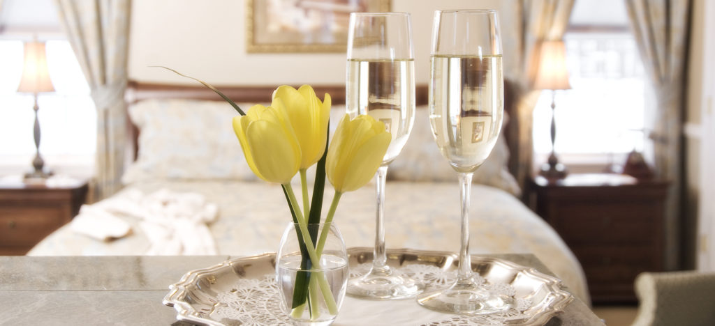 Vase with yellow tulips, champagne in crystal flutes on top of silver tray. Background faded picture of king sleigh bed with pale blue bedding with 2 windows.